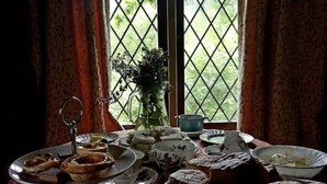 Uma mesa posta com diversos doces e sobremesas, um bule de chá e um vaso de flores.