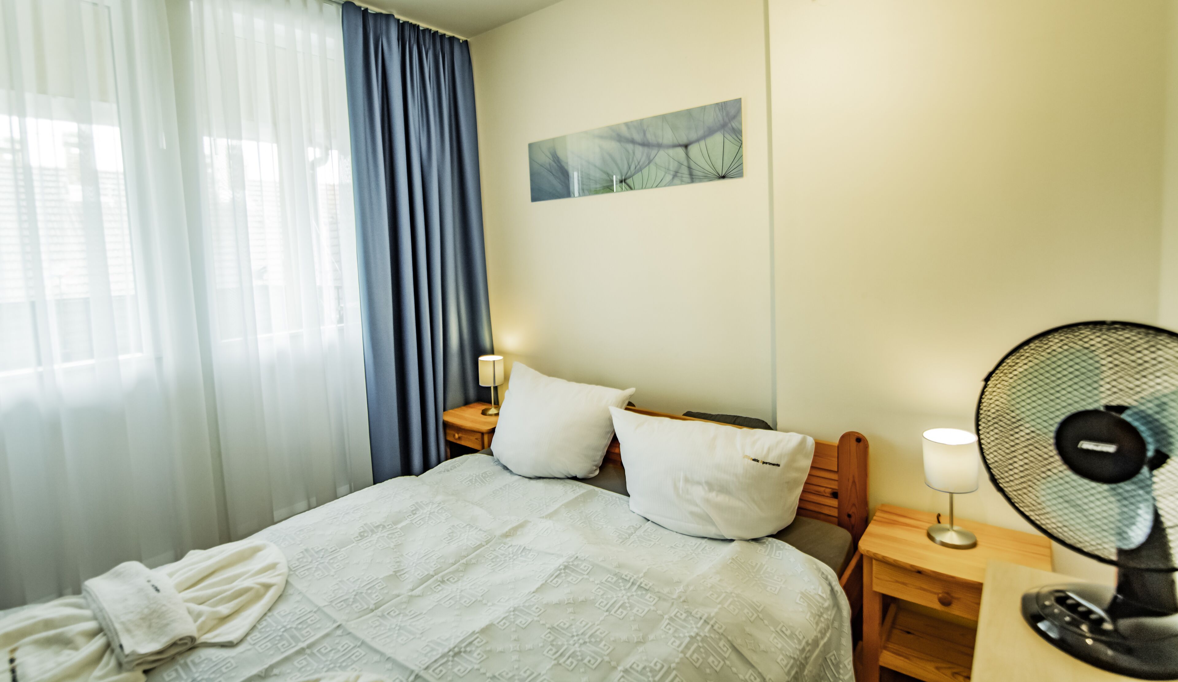 A hotel room with a bed, a nightstand, a fan, a lamp, and a window with curtains.