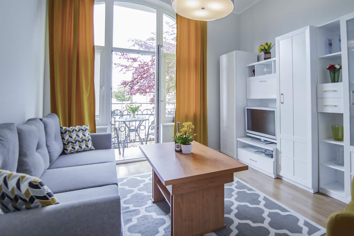 Een moderne woonkamer met een grijze bank, een houten salontafel en een gele fauteuil. Er is een televisie op een witte kast, en een balkon met gele gordijnen en een tafel met bloemen.