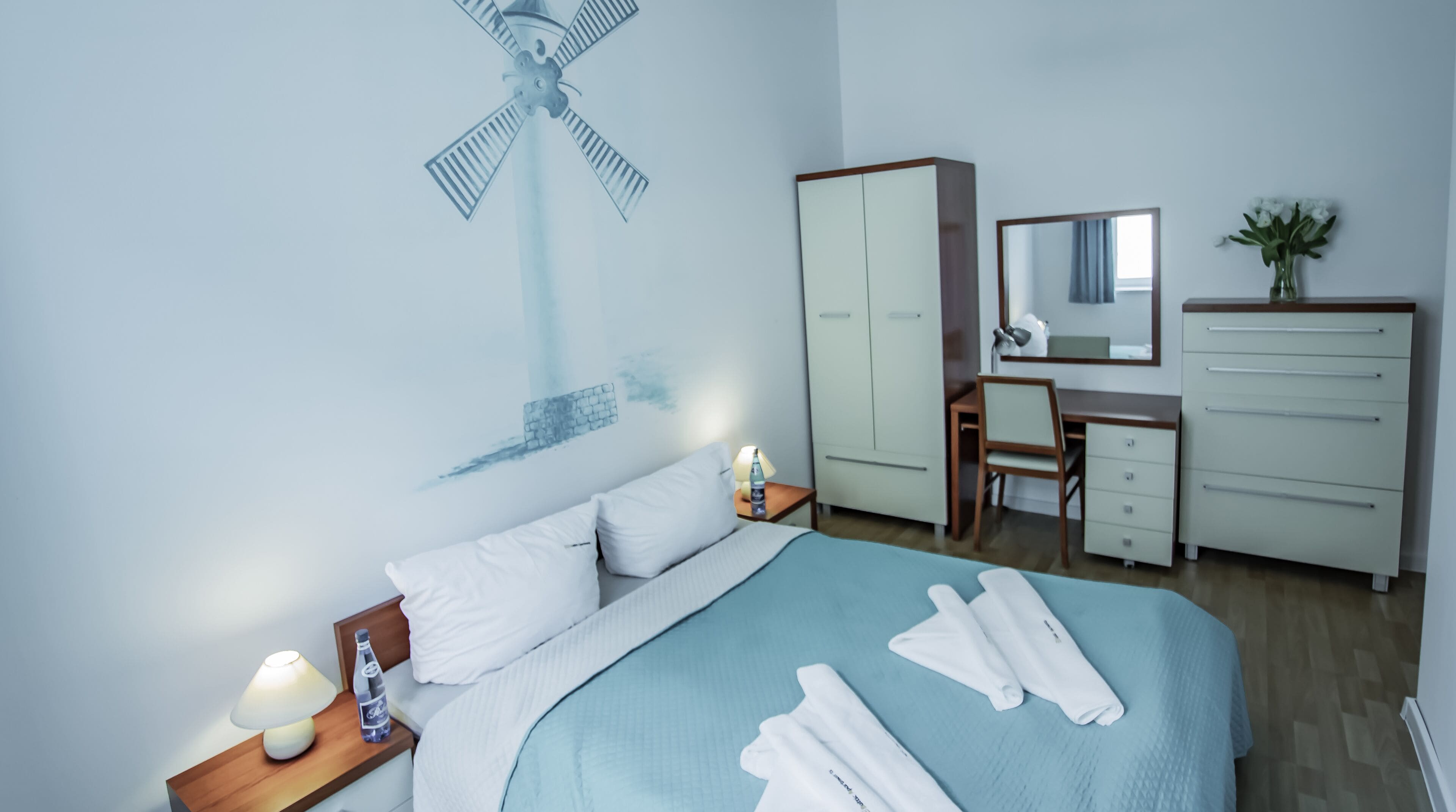 A hotel room with a bed, a desk, a wardrobe, a mirror, and a wall art of a windmill.