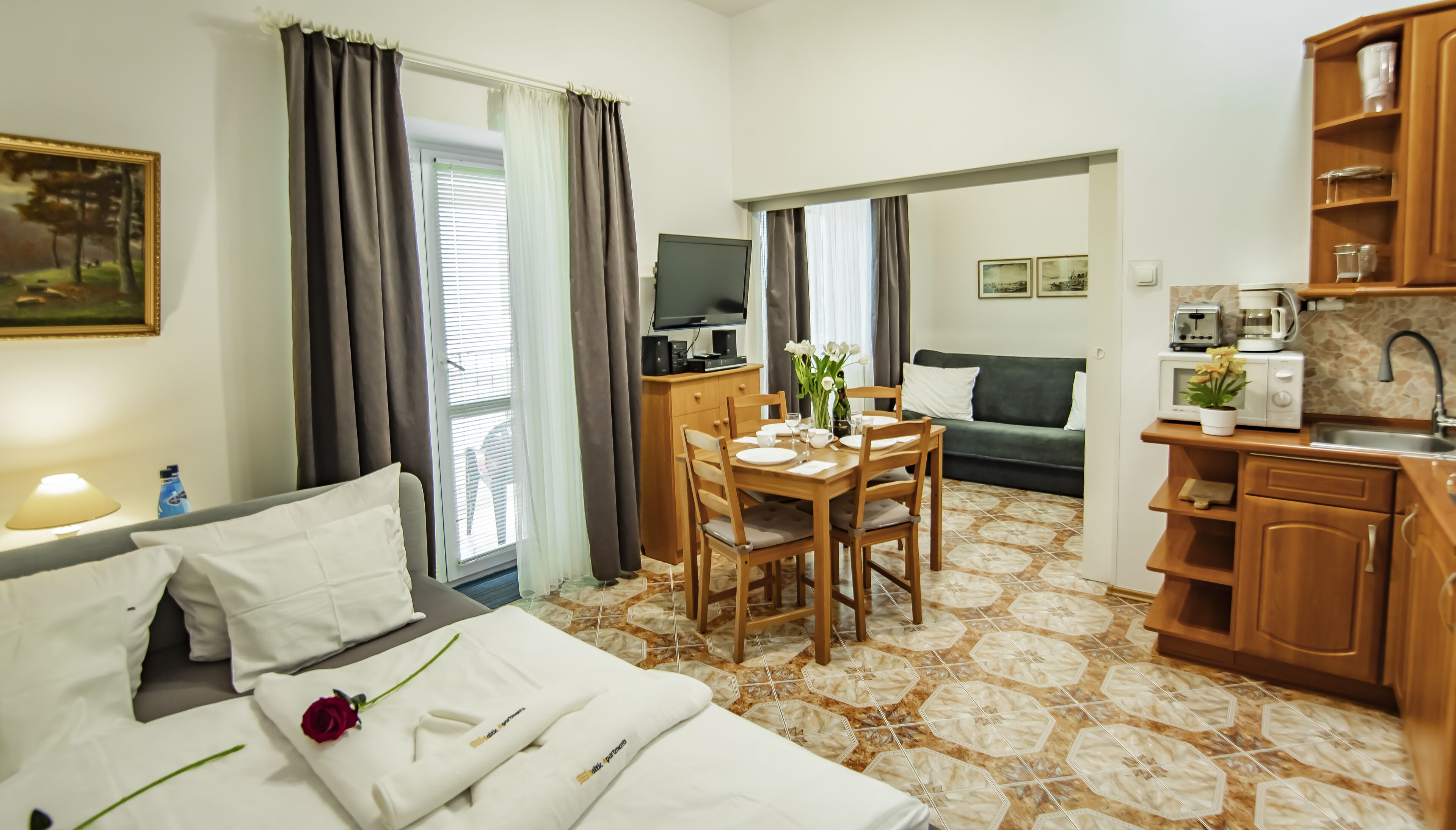 A hotel room with a bed, a dining table with chairs, a kitchenette, and a living area with a sofa.