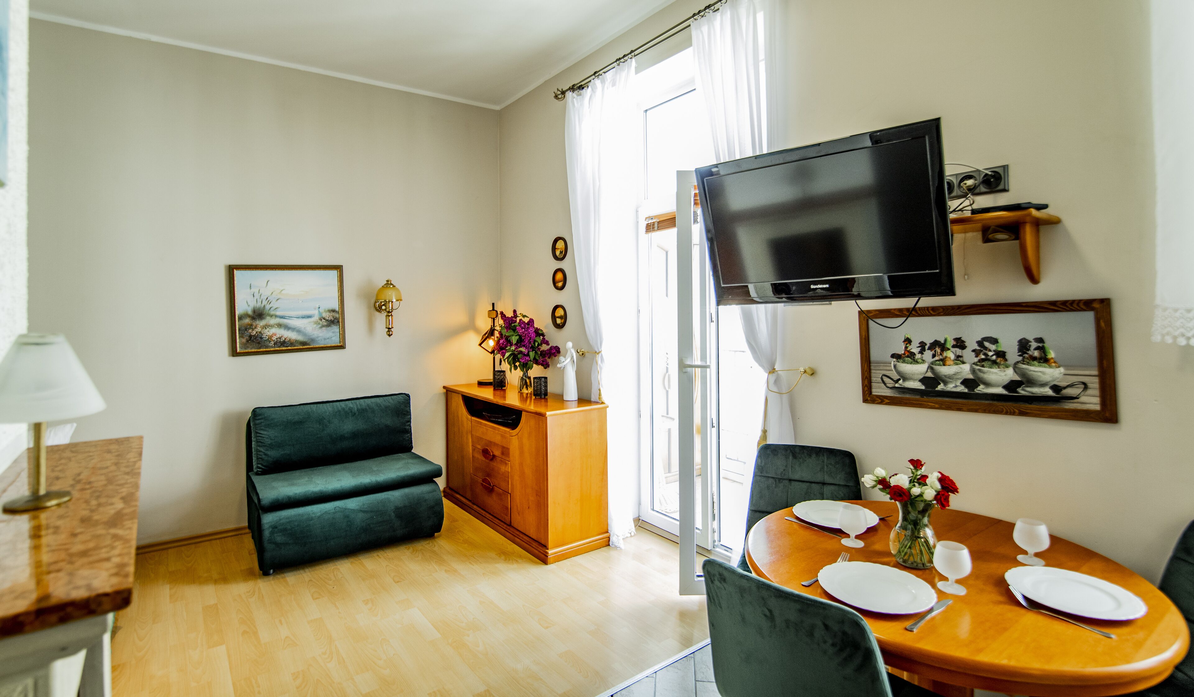 A living room with a sofa, a wooden dining table set for two, a TV mounted on the wall, and a window with curtains.