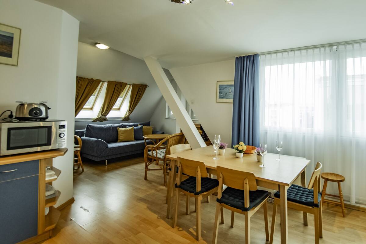 Een moderne woonkamer met een bank, een eettafel en stoelen. Er staat een magnetron op een plank, een trap en een groot raam met gordijnen.