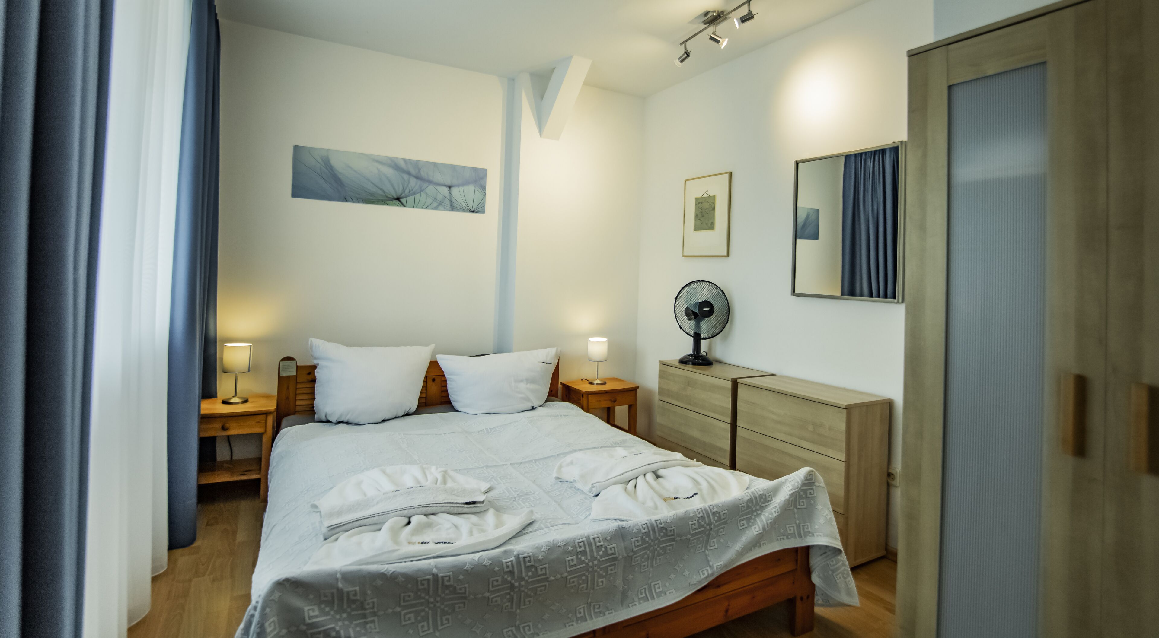 Un llit ben fet amb llençols blancs, una taula de nit amb una làmpada, un moble d'emmagatzematge i un ventilador fixat a la paret.