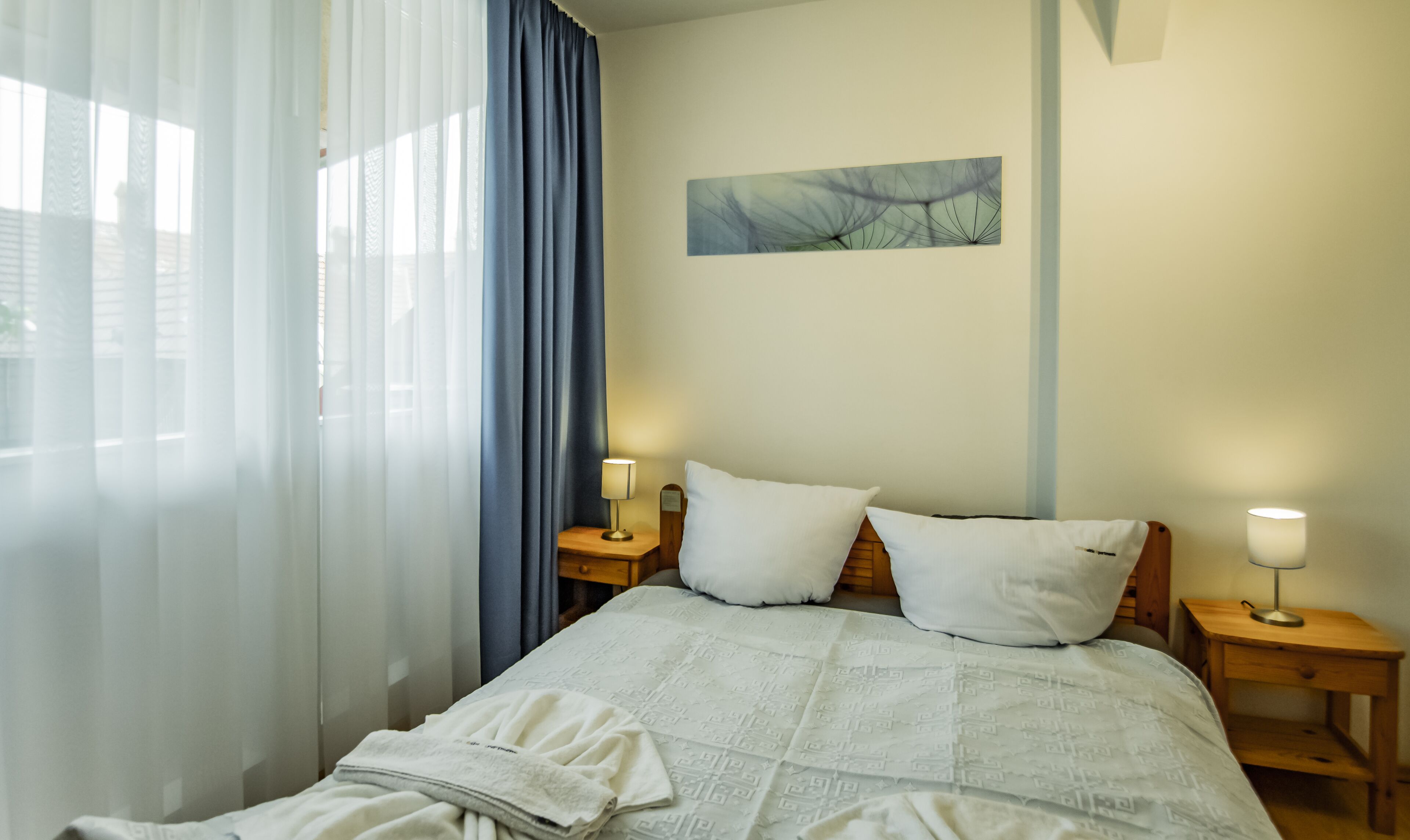 A neatly made bed with a patterned bedspread, two pillows, and a folded blanket on a wooden nightstand. A wall-mounted painting and a bedside lamp are also visible.