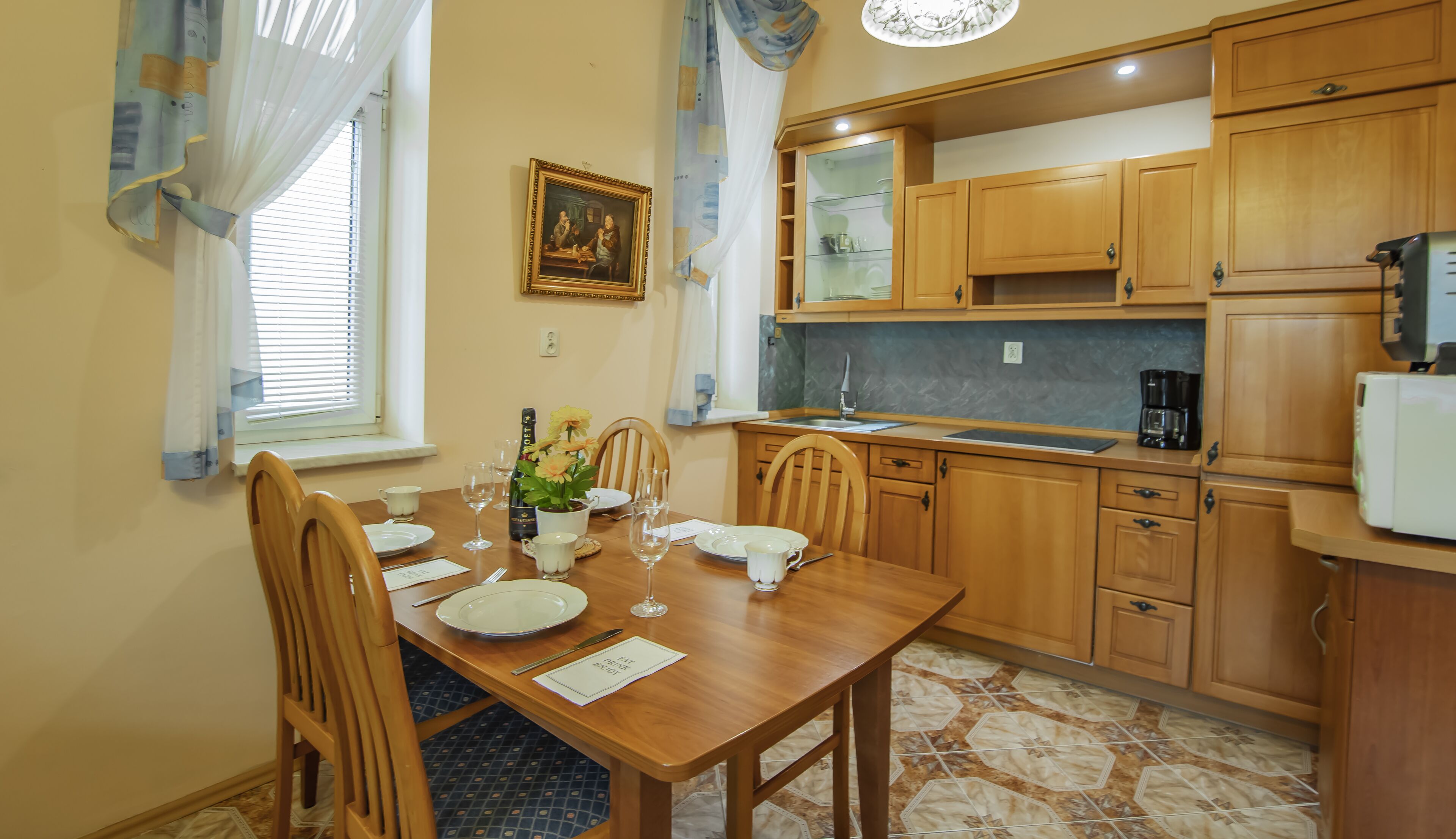 Een eethoek met een houten tafel gedekt voor vier personen, een keuken met houten kastjes en een raam met jaloezieën.
