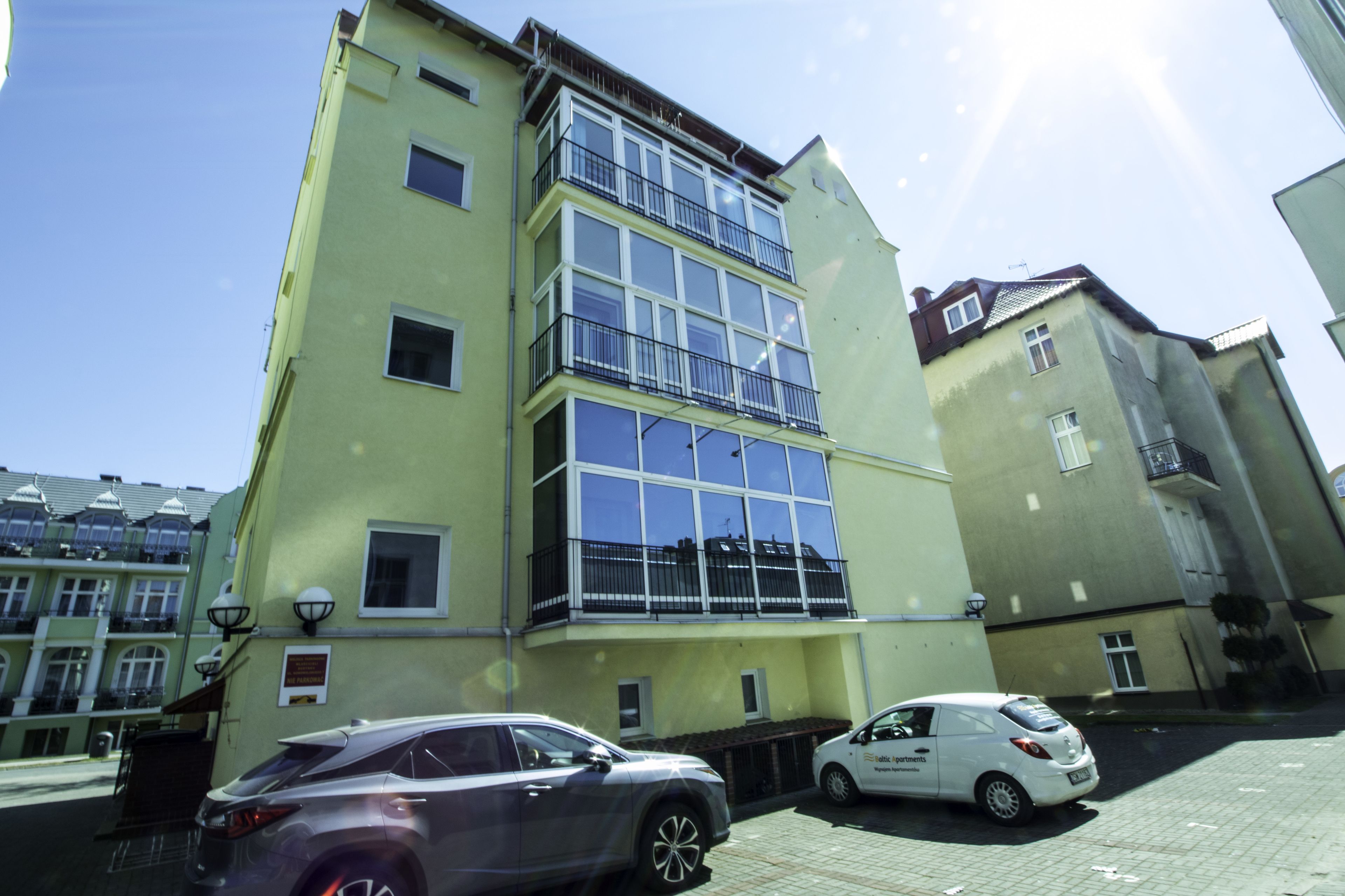 A multi-story building with large windows and balconies, parked cars in front, and a clear sky.