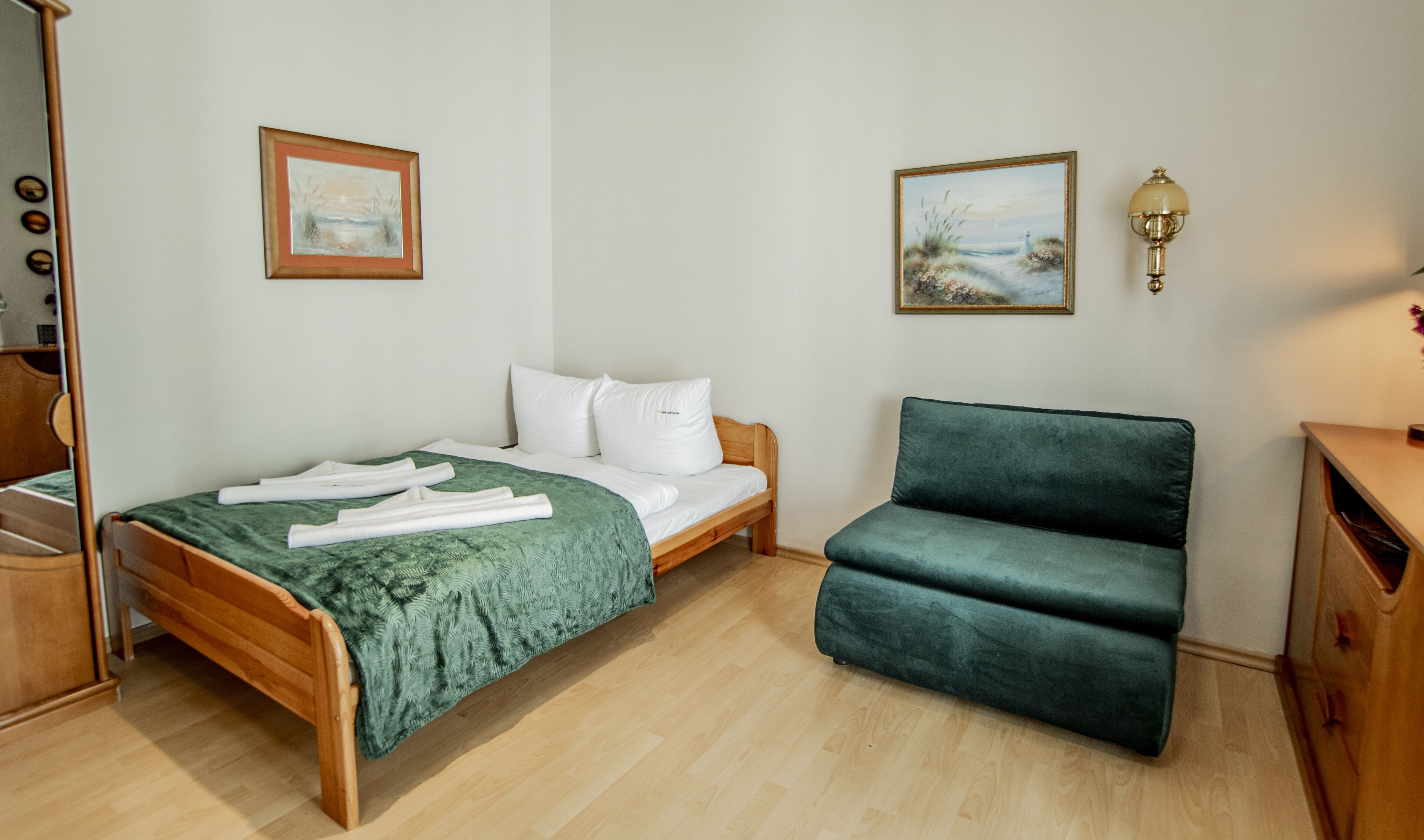 A neatly arranged bedroom with a bed, a green chair, and two framed pictures on the wall.