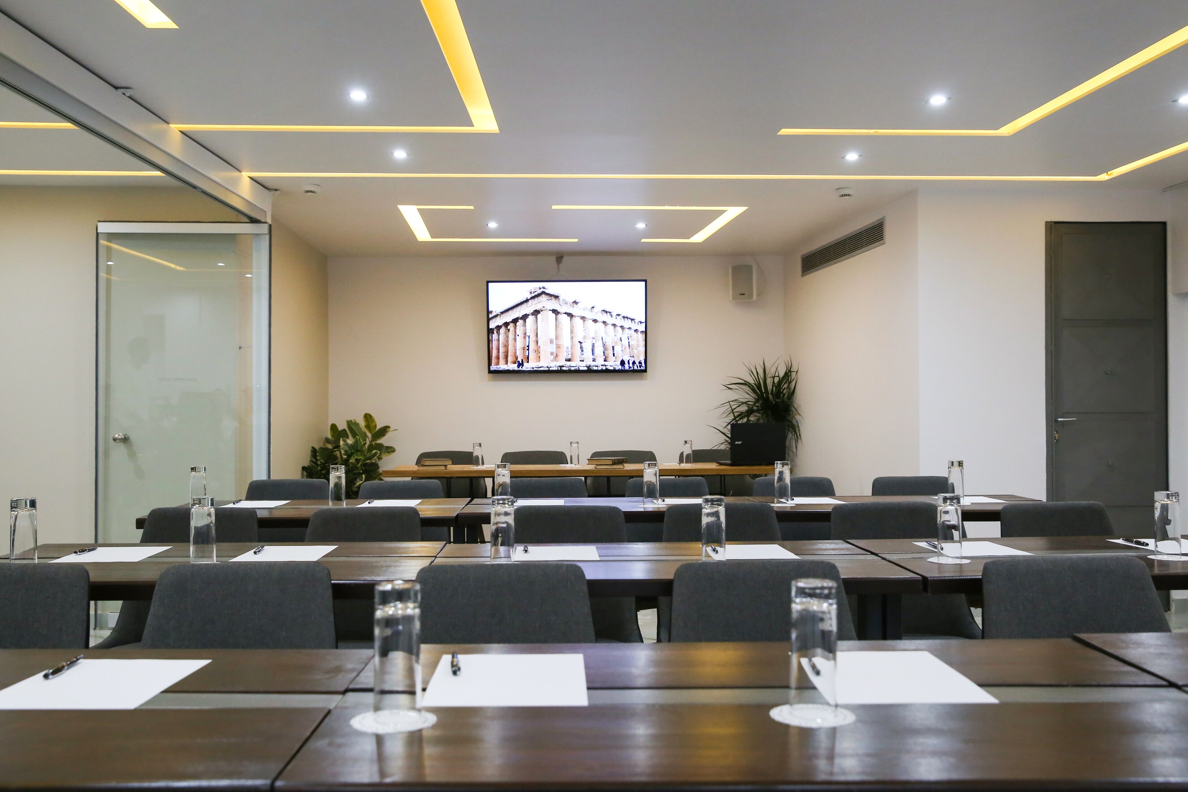 A conference room with a large screen displaying a building, multiple tables with chairs, and a glass door on the left.