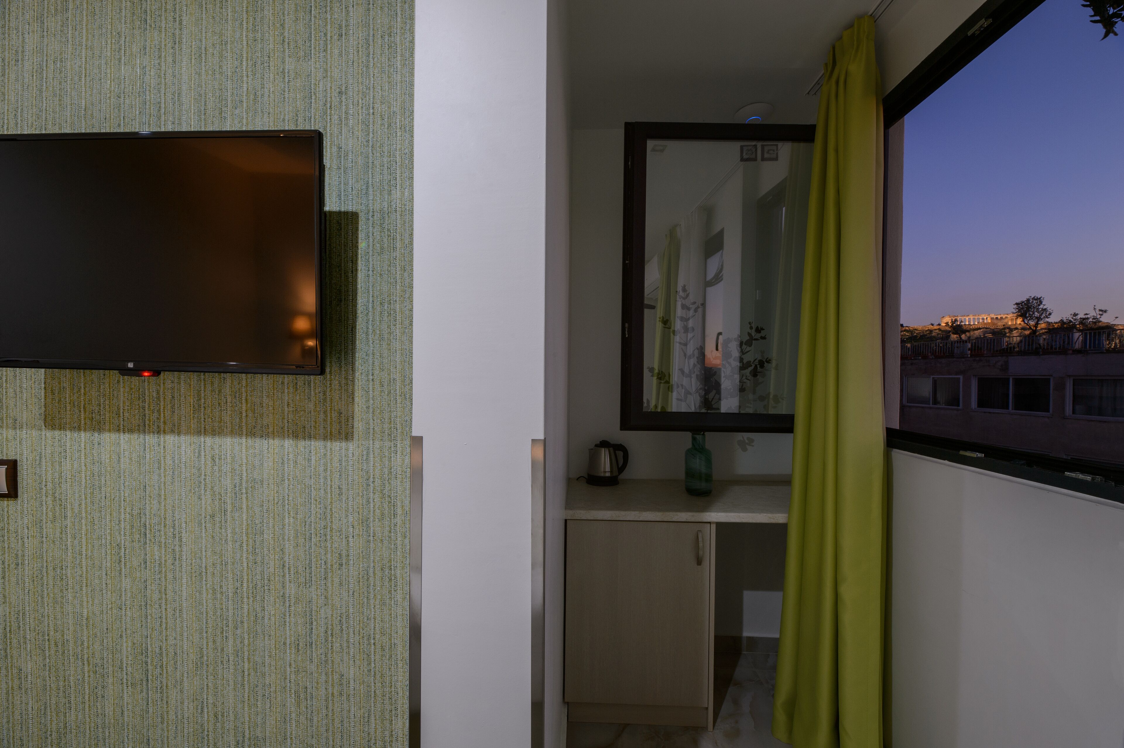 Et hotelværelse med et fladskærms-tv på væggen, et lille skab, et spejl med dekorativ ramme og udsigt til en bygning gennem et vindue med grøn gardin.