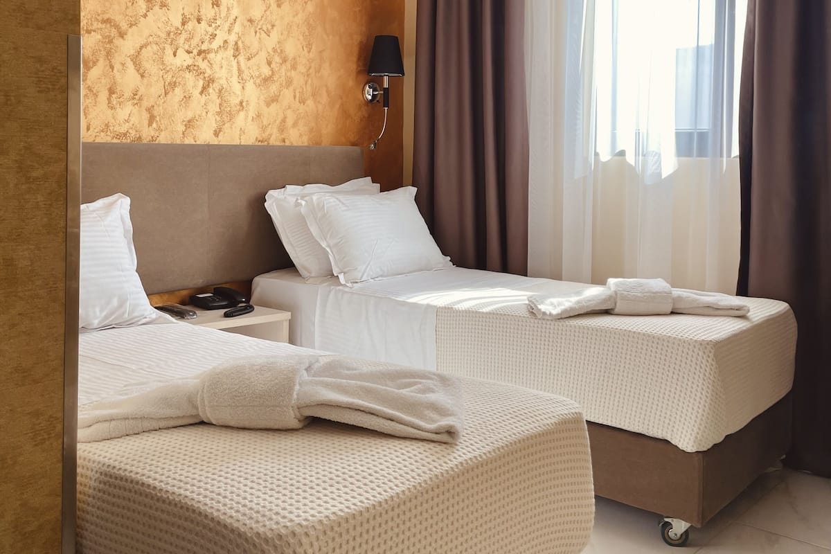 A hotel room with two beds, a wall-mounted lamp, and a window with curtains.