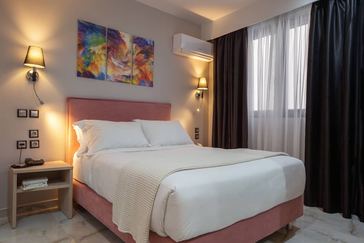 A hotel room with a large bed, bedside lamps, a nightstand, a wall-mounted air conditioner, and a window with curtains.