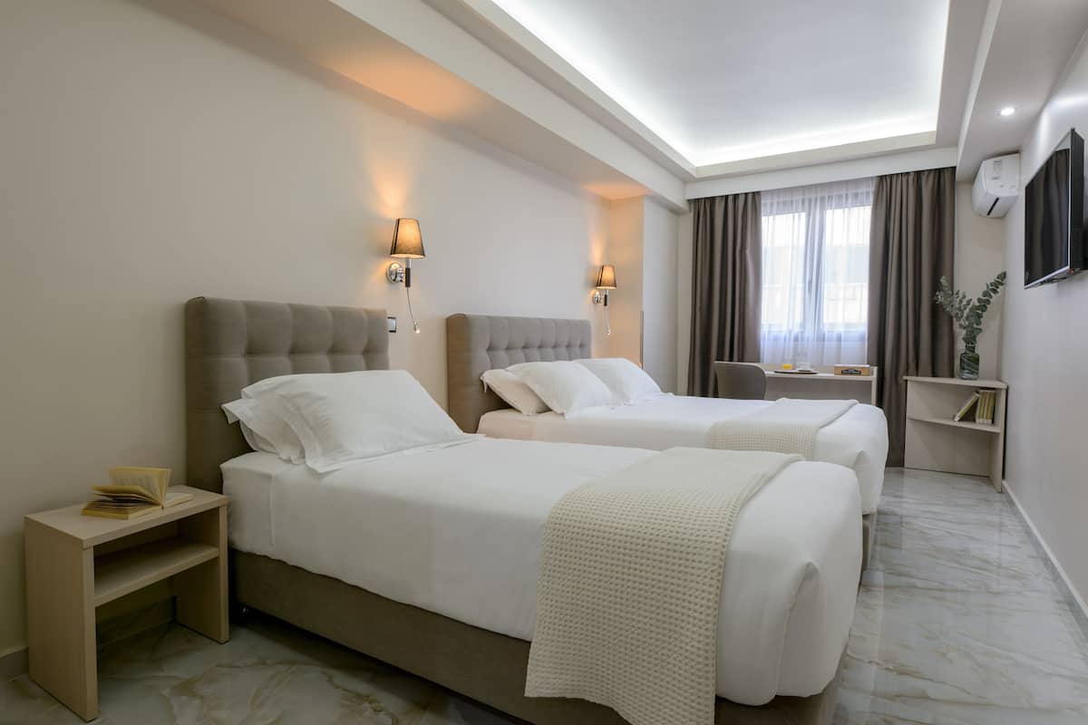A hotel room with two beds, a bedside table with a lamp, a small desk, a TV mounted on the wall, and a window with curtains.