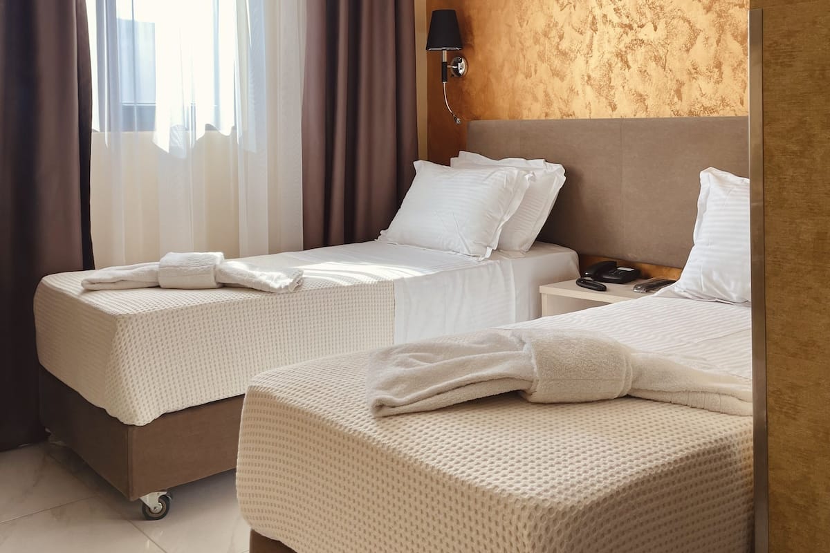 A hotel room with two beds, a nightstand, a lamp, and a view of the city through sheer curtains.