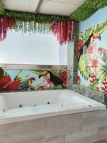 Casa de banho com uma banheira grande, um mural na parede com aves e flores tropicais, e plantas pendentes.