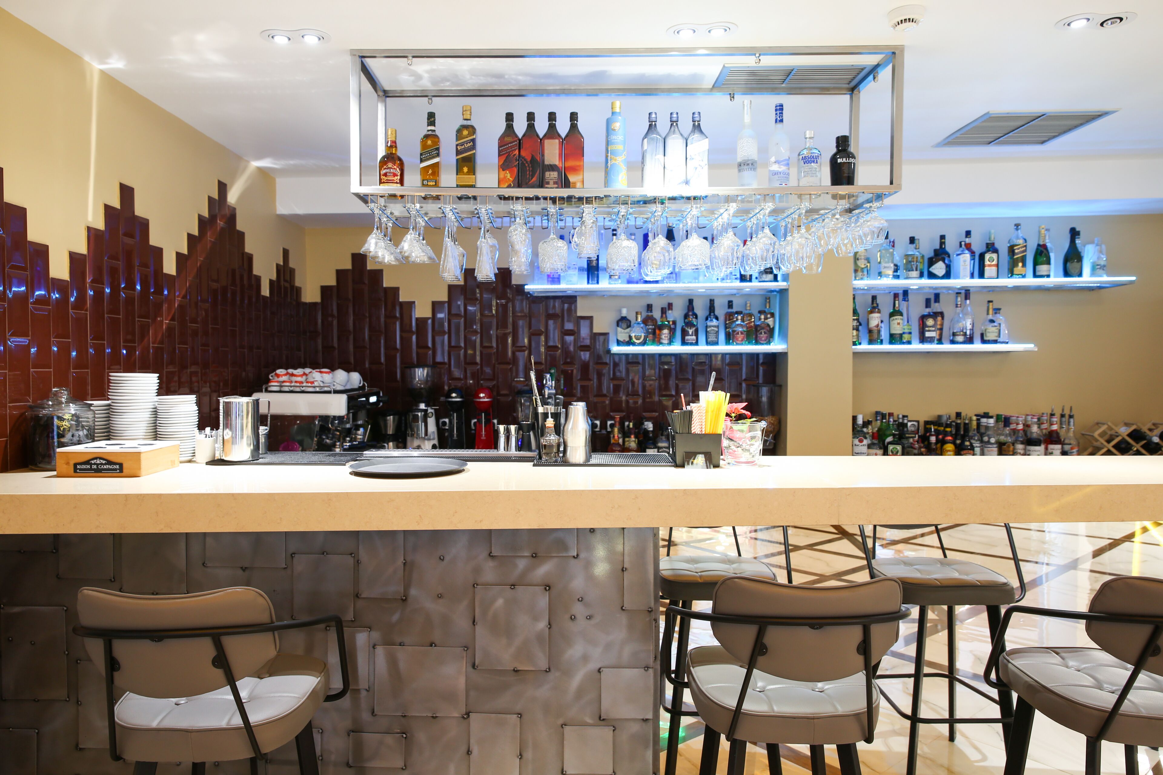 A bar area with a counter, shelves of liquor bottles, hanging glasses, and bar stools.