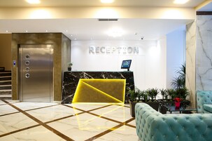 Uma área de recepção com piso de mármore, balcão de recepção, placa indicativa, tela digital e escada.