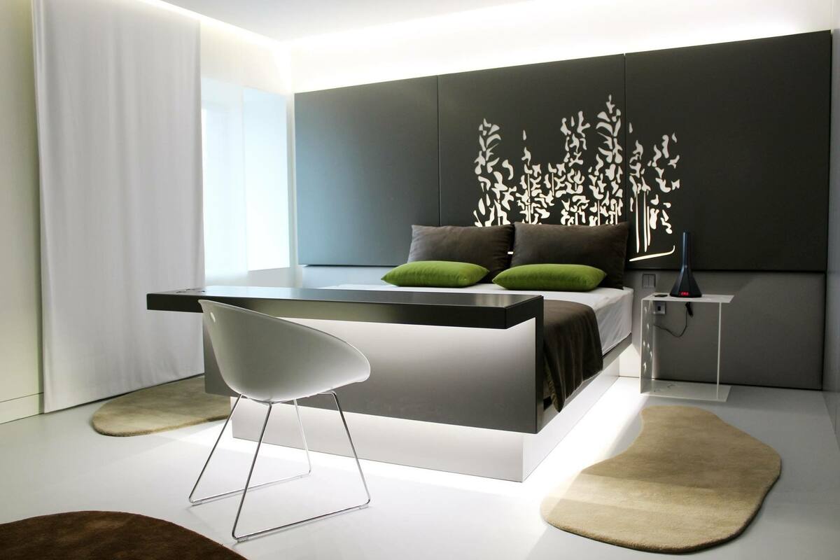 A modern bedroom with a bed, bedside table, chair, and decorative wall panel.