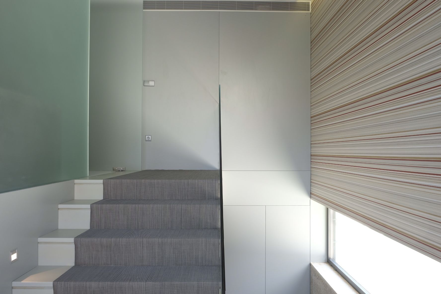 Una escalera con peldaños alfombrados de color gris y paredes blancas, que conduce a una habitación con un gran ventanal y persianas.