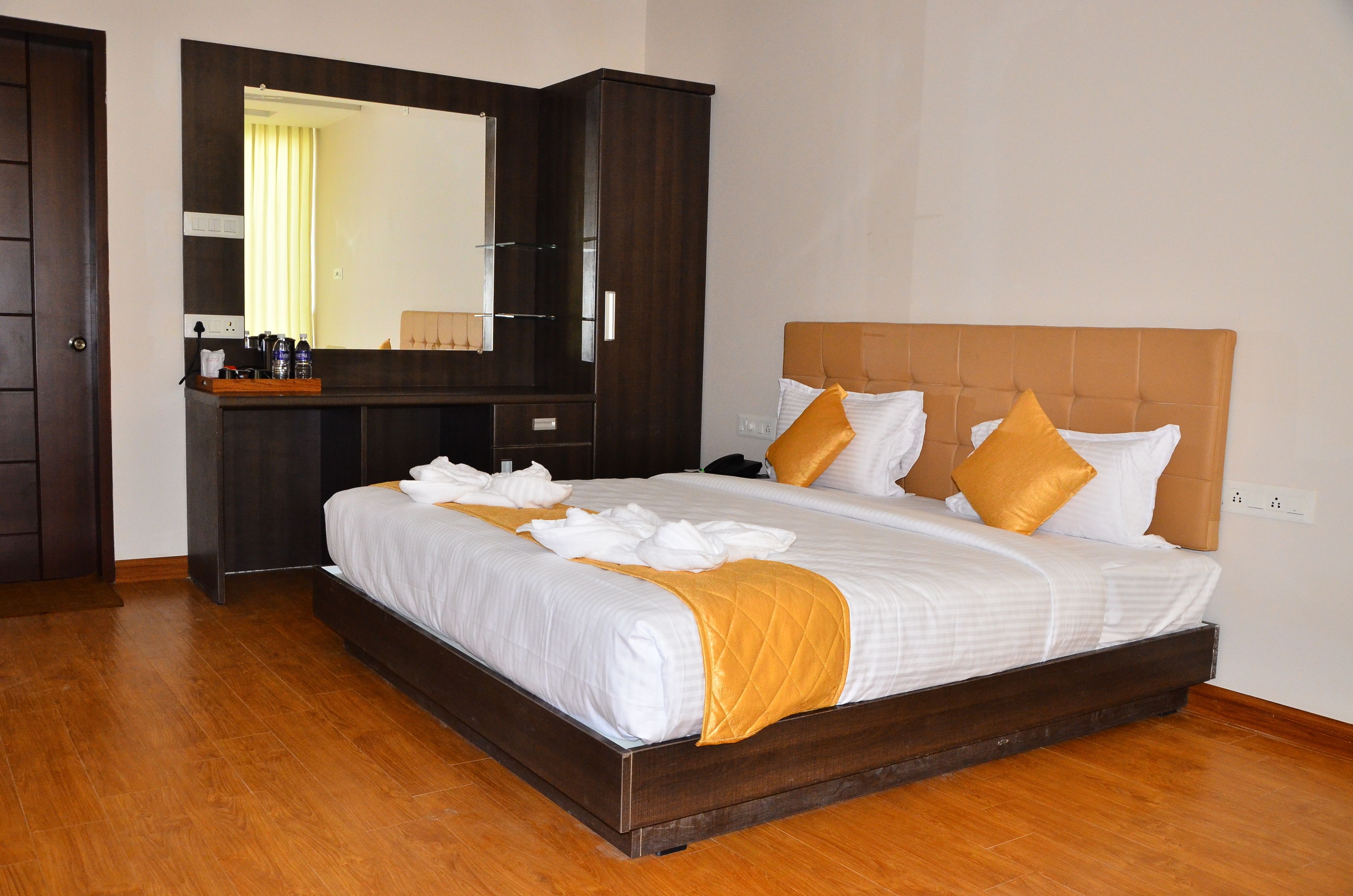 A hotel room with a large bed, a wooden headboard, a dark wooden wardrobe, a mirror, and a small desk with toiletries.