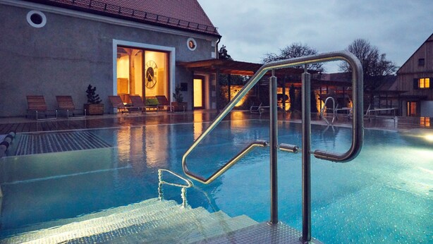 A swimming pool with a metal ladder, lounge chairs, and a building with a tiled roof in the background. - Romantik Hotel Zum Klosterbräu (Neuburg)