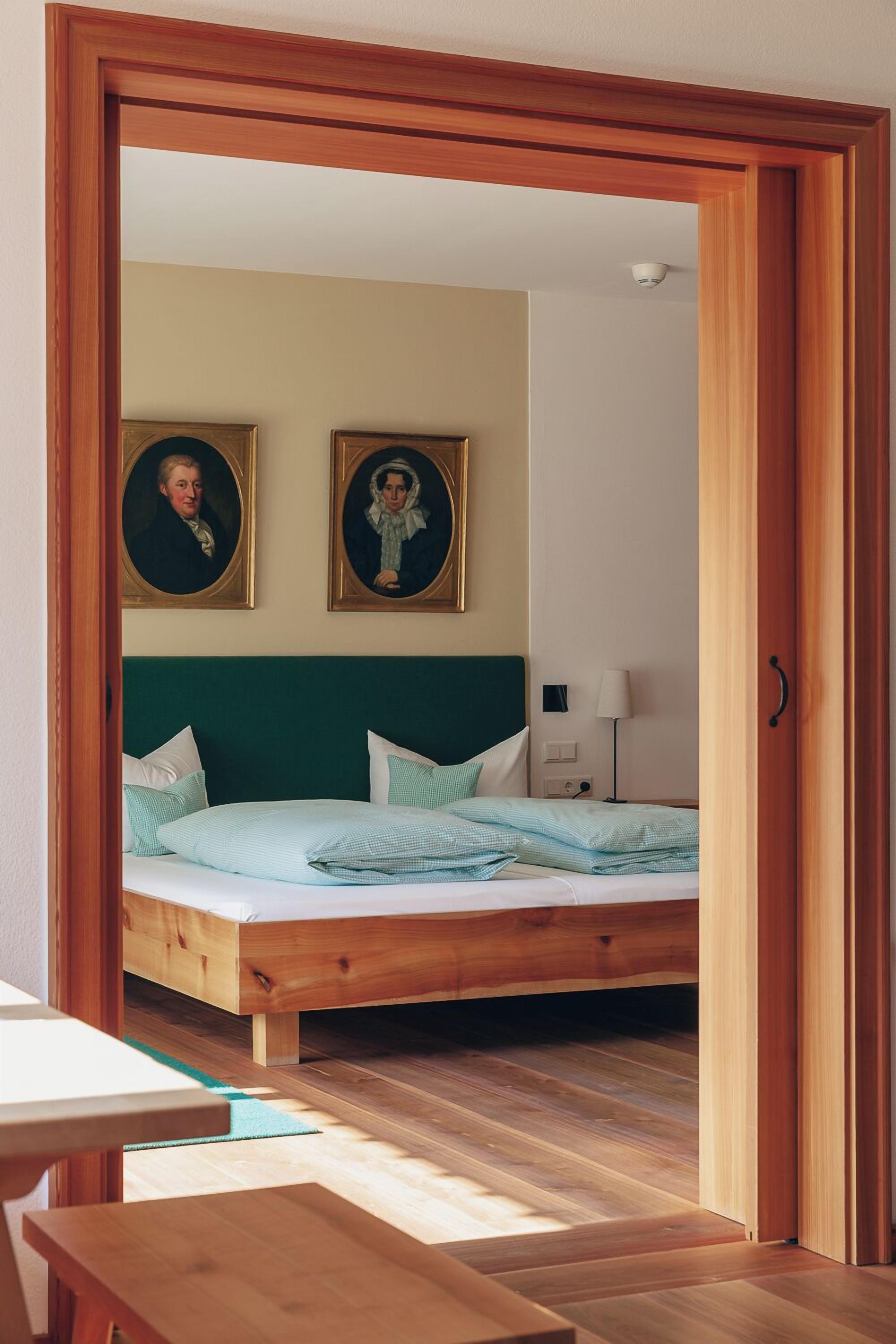 A room with a wooden bed, a green headboard, two framed portraits on the wall, and a bedside table.