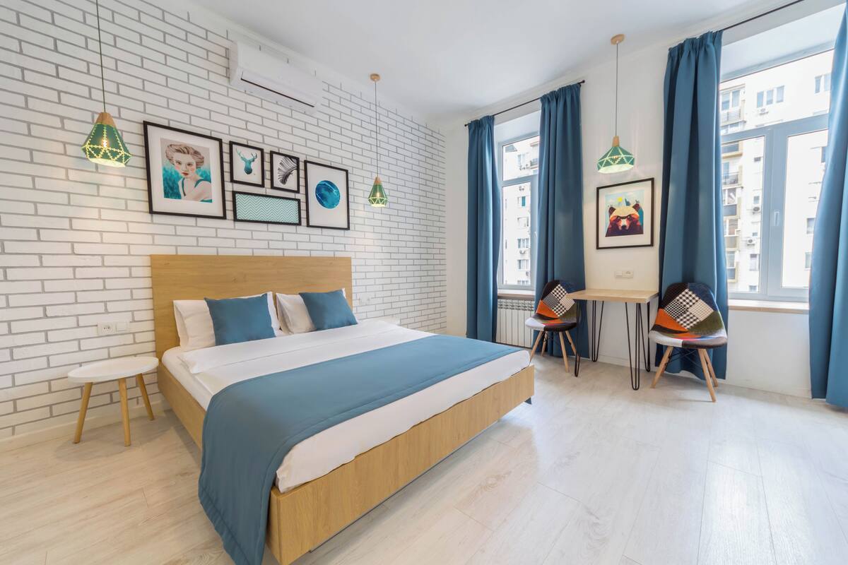 A modern hotel room with a wooden bed, a small desk with chairs, a white brick wall, and framed pictures.