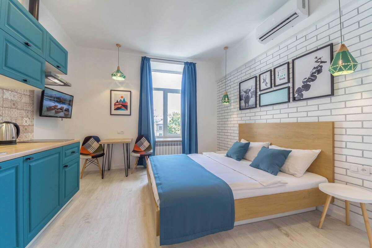 A modern bedroom with a wooden headboard, a large bed with blue and white bedding, a small bedside table, and a view of the city through a window with blue curtains.