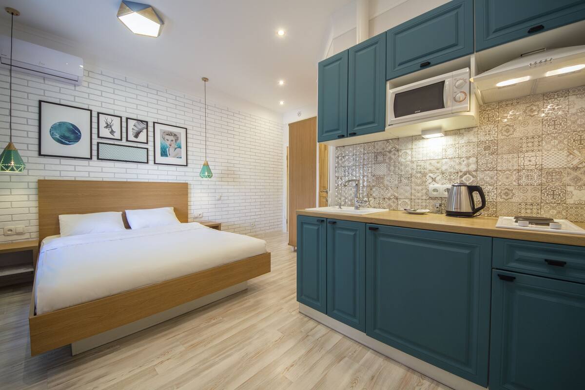 A modern bedroom with a wooden bed and a kitchenette with teal cabinets and a microwave.