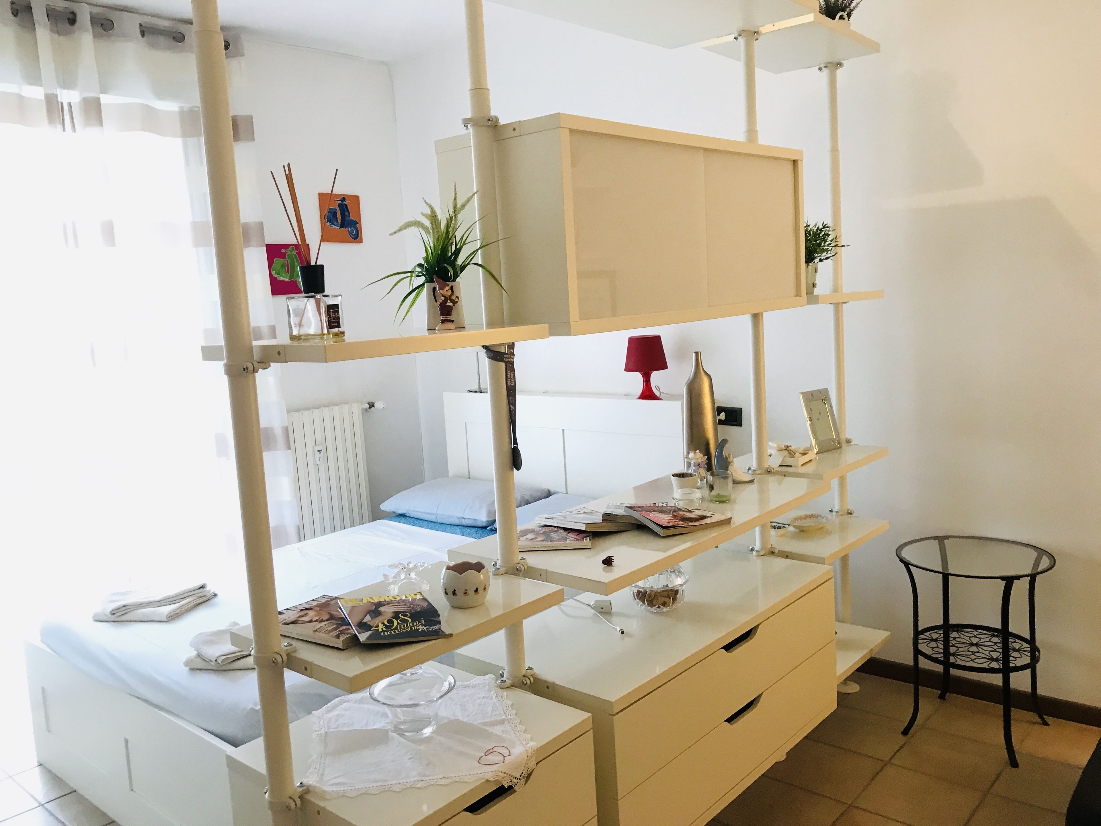 Een netjes opgeruimde slaapkamer met een bed, nachtkastjes, een klein rond tafeltje en een wandplank met diverse spullen.