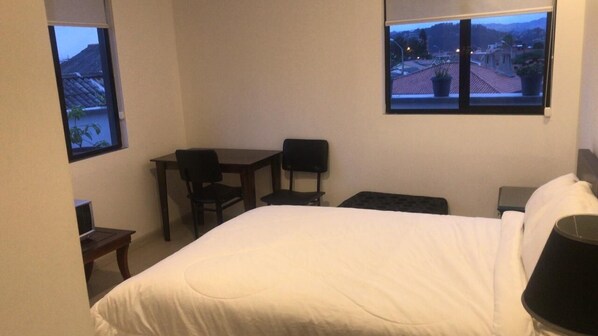 A hotel room with a bed, a desk, two chairs, a small table, and two windows offering views of a town and mountains.