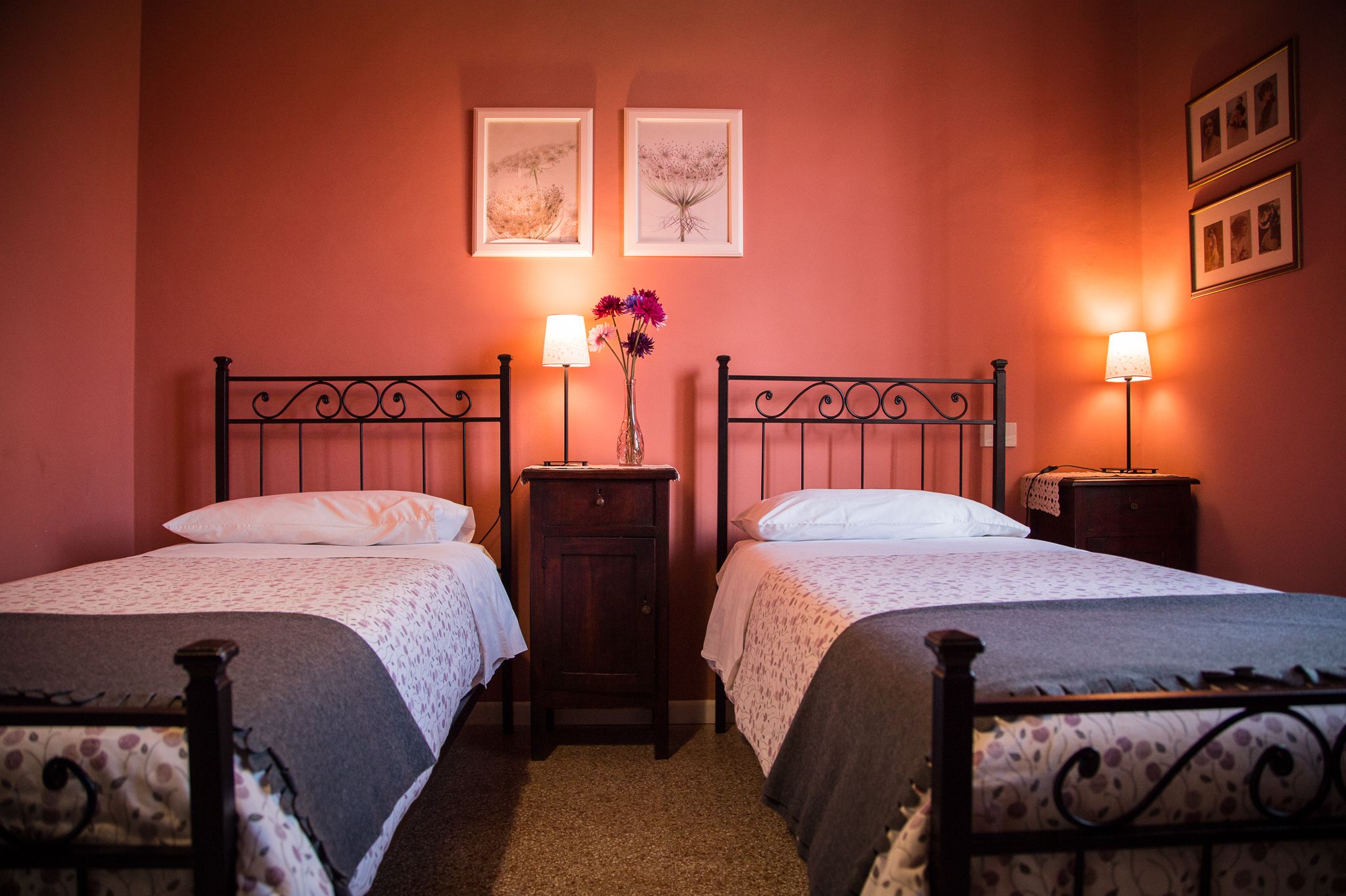 A room with two single beds, a nightstand with a lamp, a vase with flowers, and two framed pictures on the wall.