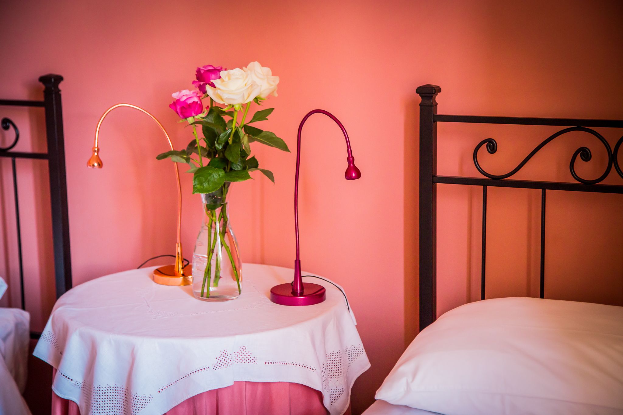 Una cama con un almohadón blanco, una mesa redonda con mantel blanco y un florero con flores.