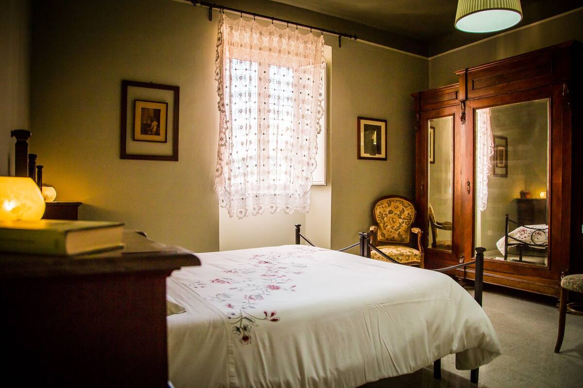 A bedroom with a bed, a chair, a wardrobe, and a window with lace curtains.