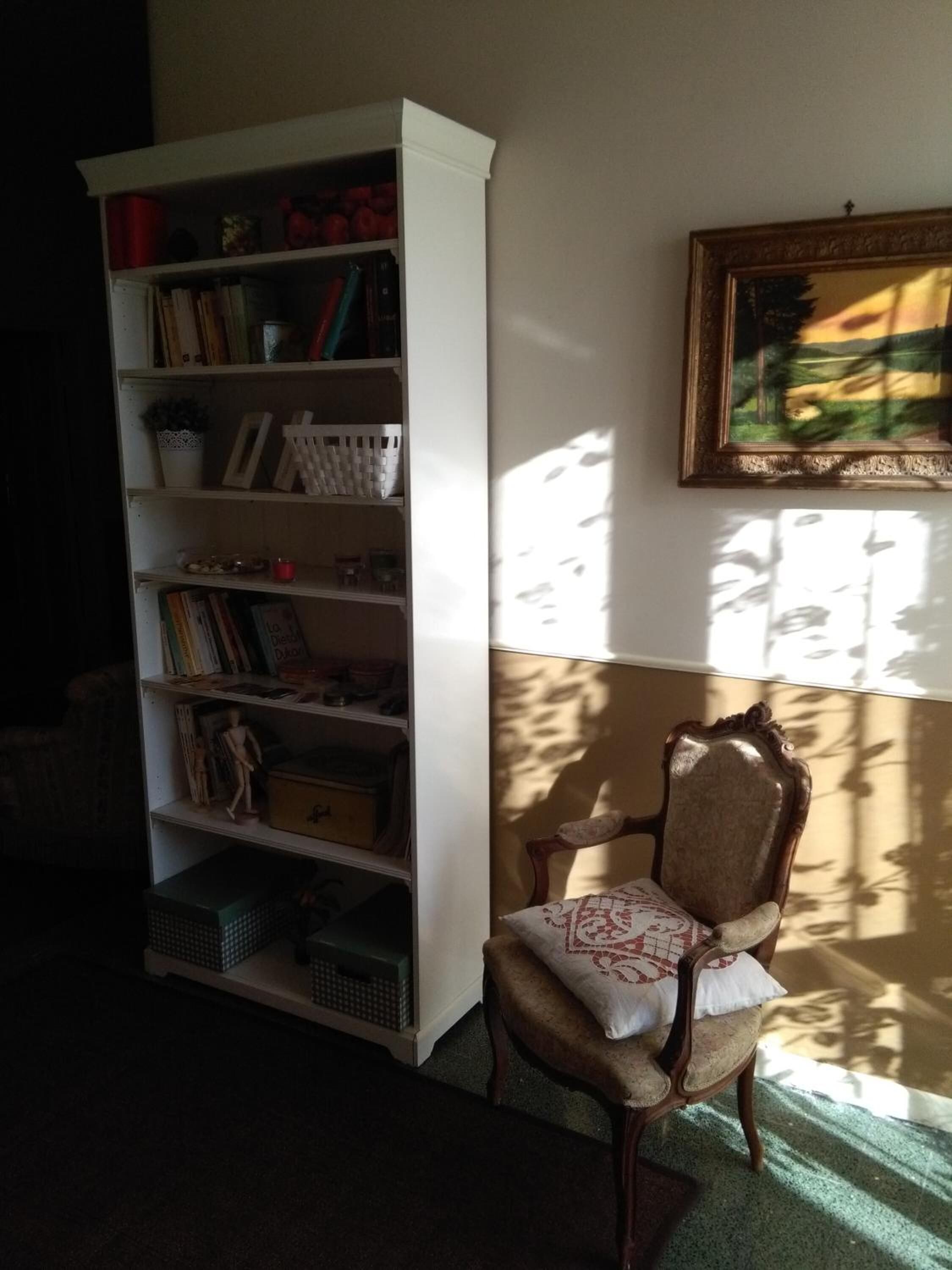 A room with a white bookshelf filled with books and decorative items, a wooden chair with a cushion, and a painting on the wall.
