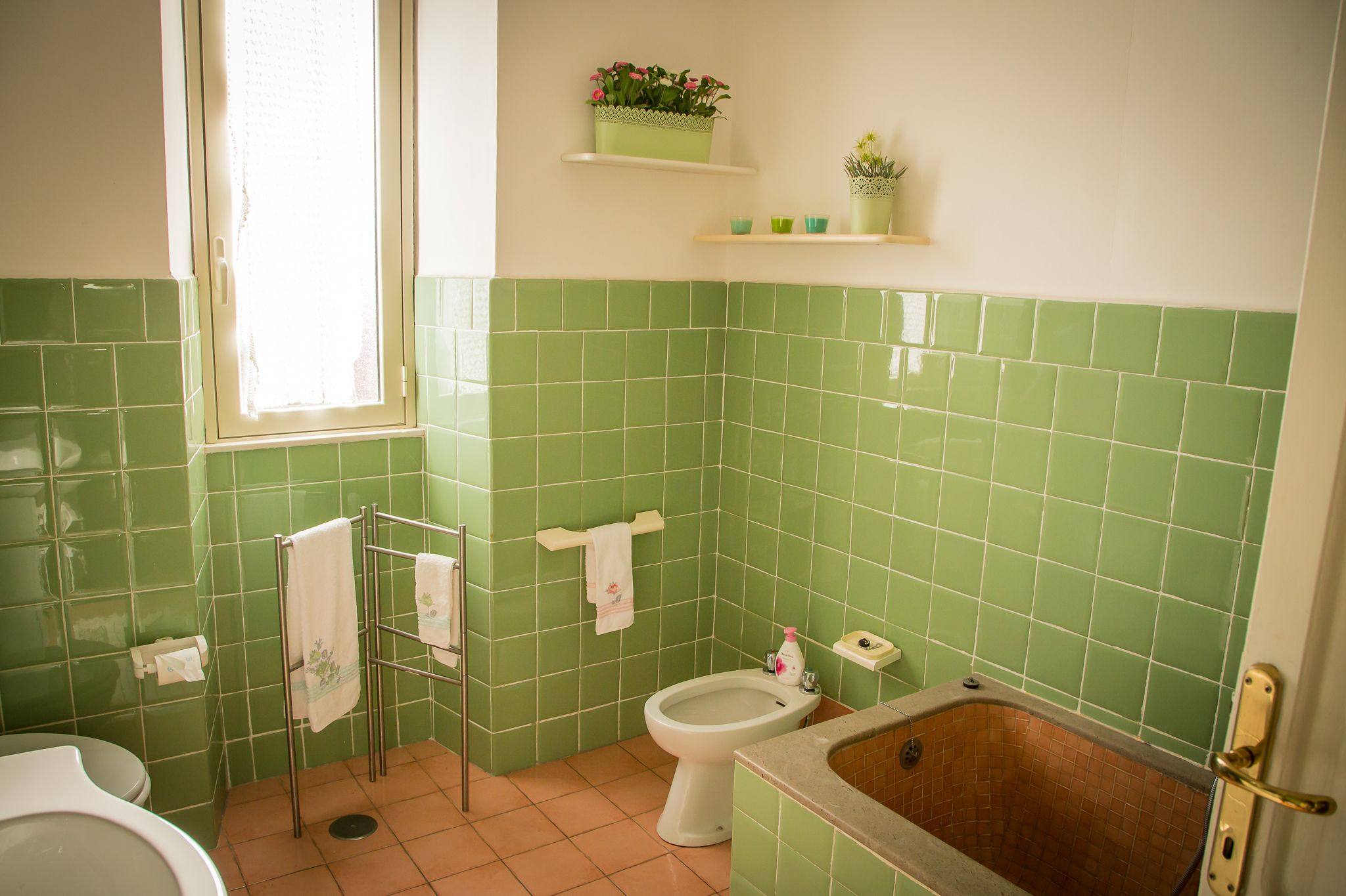 A bathroom with green tiles, a toilet, a bidet, a bathtub, and a window with a curtain.
