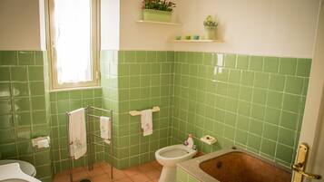 A bathroom with green tiles, a toilet, a bidet, a bathtub, and a window with a curtain.