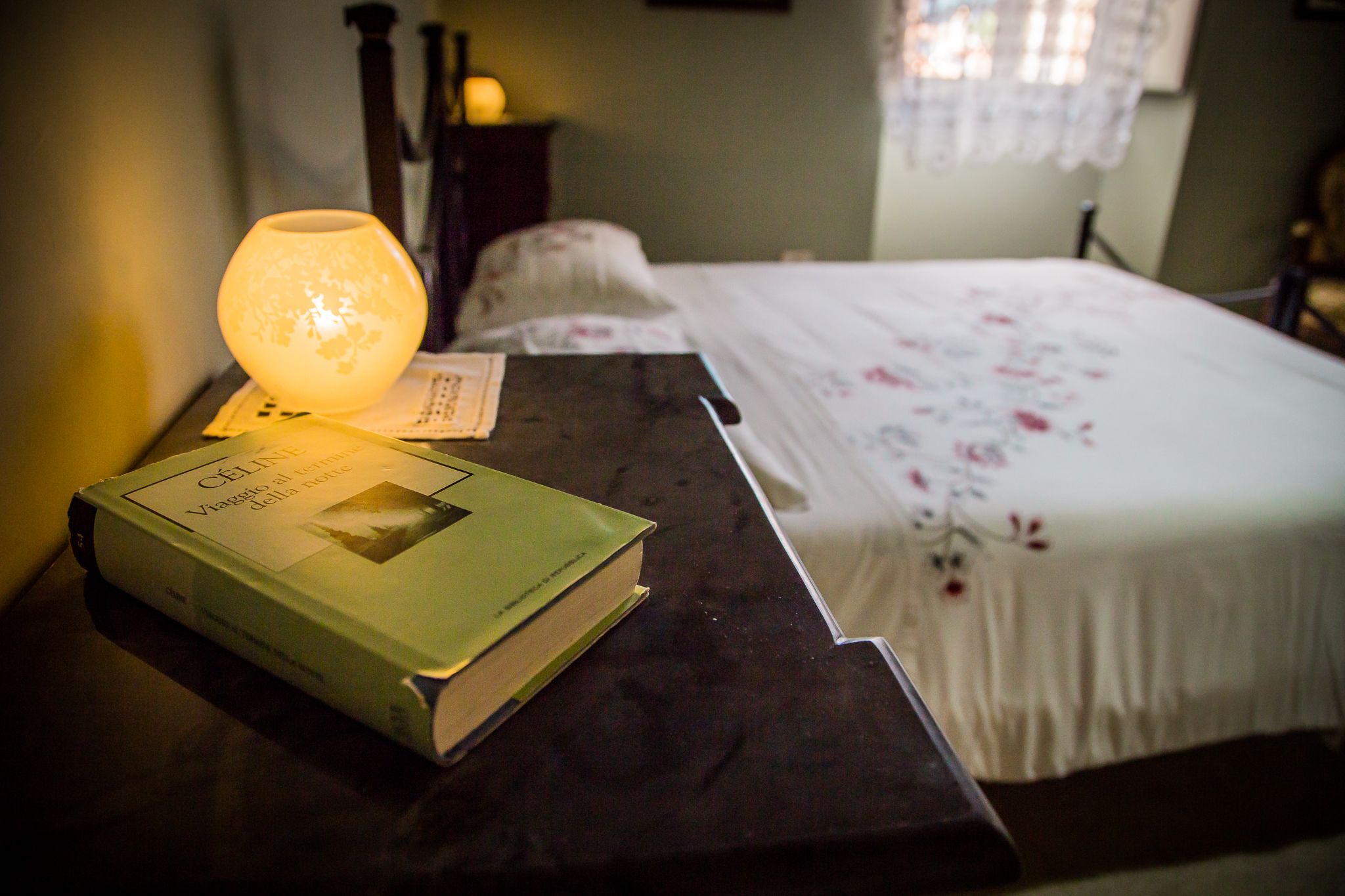 Un lit recouvert d’une couvre-lit à motifs floraux, une table de chevet avec un livre et une lampe allumée, et une fenêtre avec des rideaux.