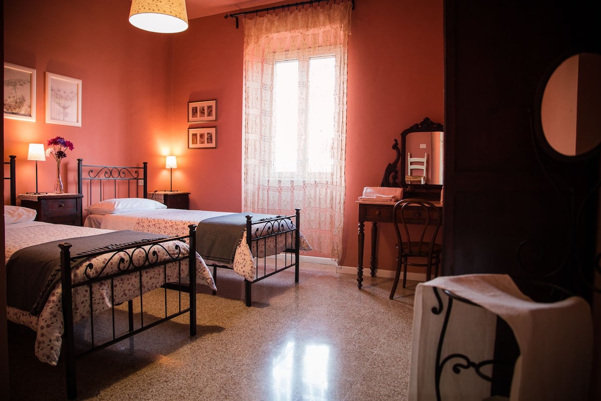 A bedroom with two beds, a desk, a chair, a mirror, and a window with lace curtains.