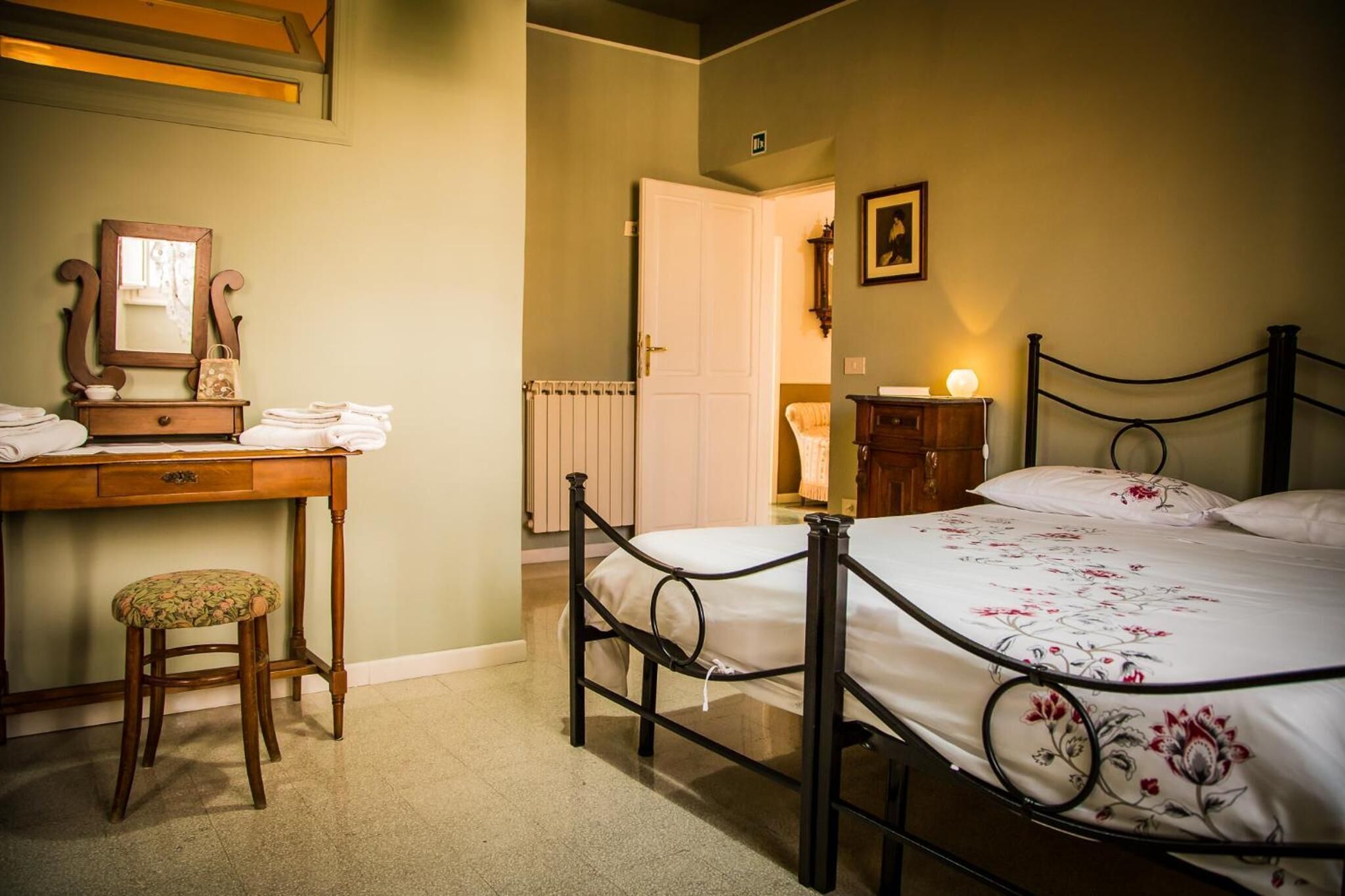 A bedroom with a bed, a desk with a chair, a radiator, and a door leading to another room.