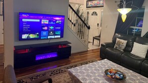 A living room with a flat-screen TV mounted on the wall, a black leather sofa, a patterned tablecloth, and a decorative bowl on the table.