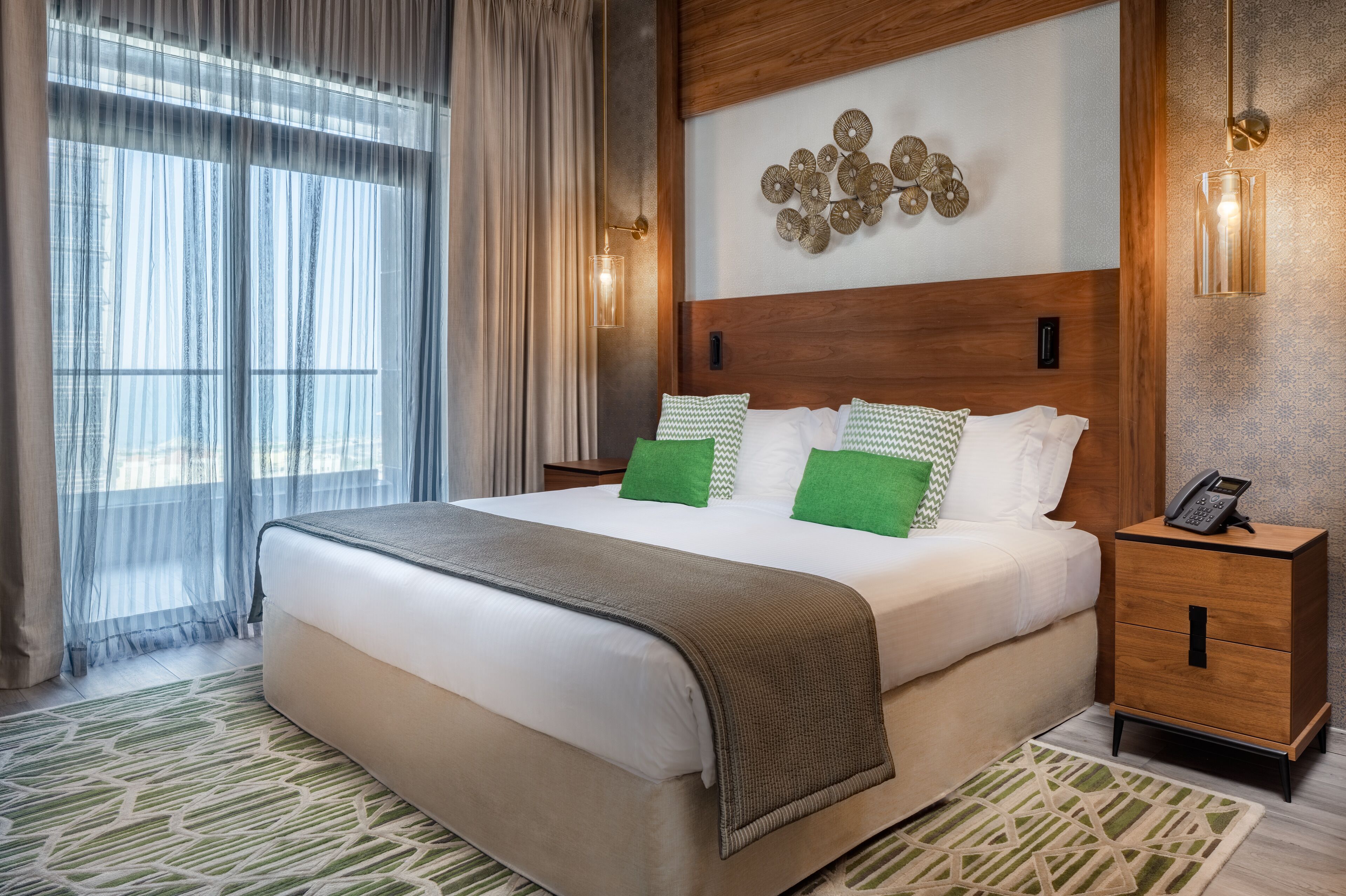 A hotel room with a large bed, a wooden headboard, a bedside table with a phone, and a decorative wall piece.