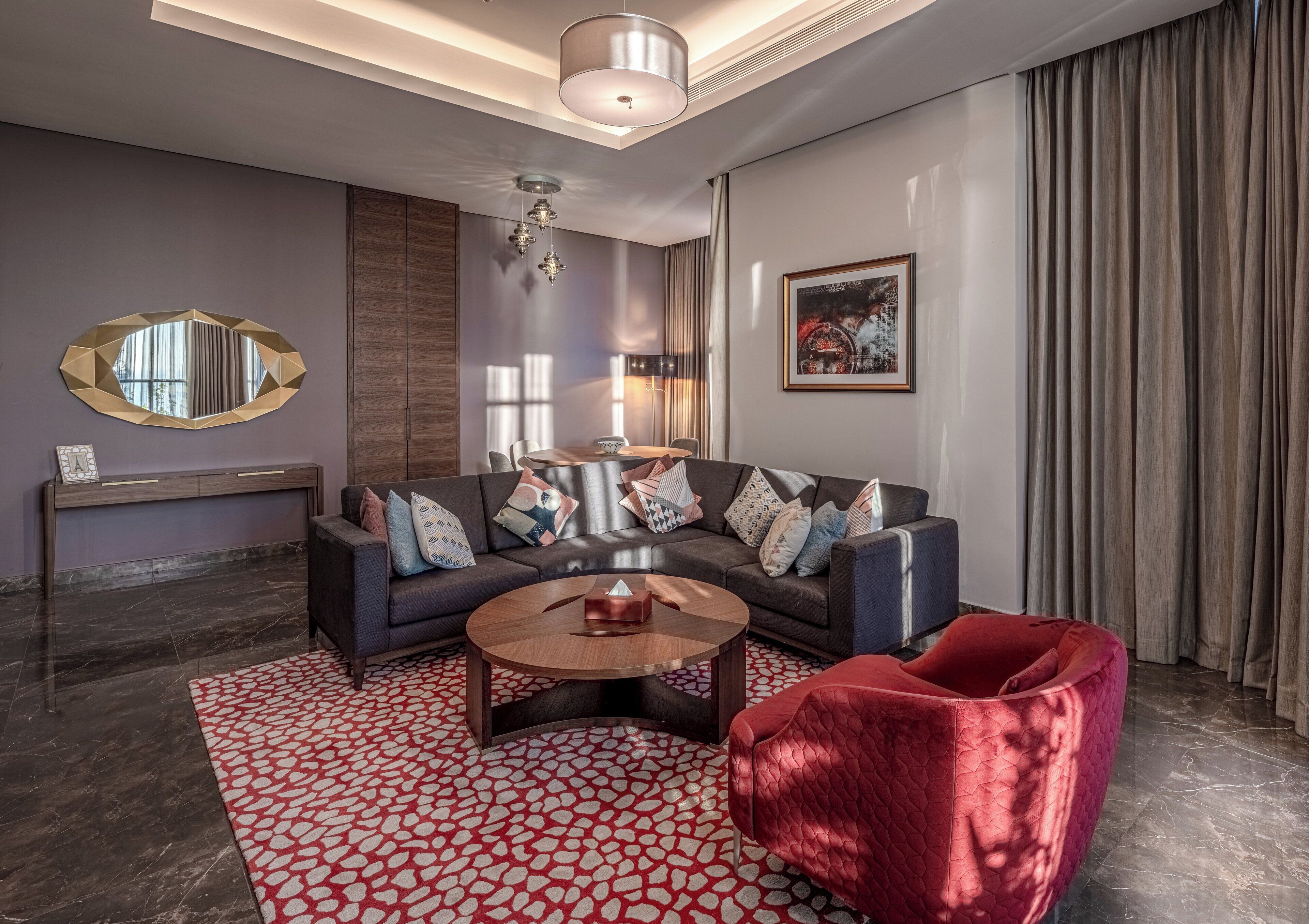 Una sala de estar moderna con un sofá gris oscuro, un sillón de terciopelo rojo, una mesa de centro redonda y una alfombra estampada.