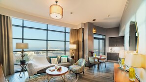 Quarto de hotel com uma grande janela, sofá, cadeira, mesa pequena e escrivaninha com luminária.