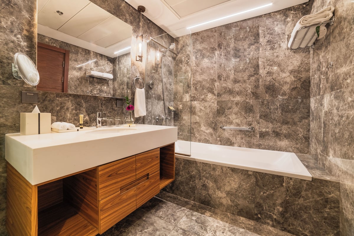 Un baño moderno con pared de mármol, tocador de madera, bañera blanca y mampara de vidrio.