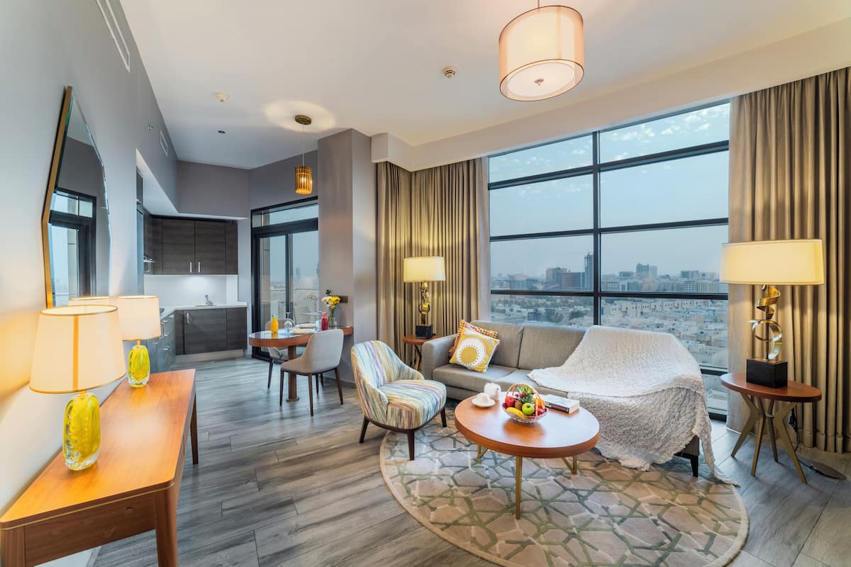 Habitación de hotel moderna con un gran ventanal, sofá, zona de comedor y cocina al fondo.