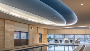 Piscina interna com design moderno, rodeada por espreguiçadeiras e vista para a paisagem urbana.