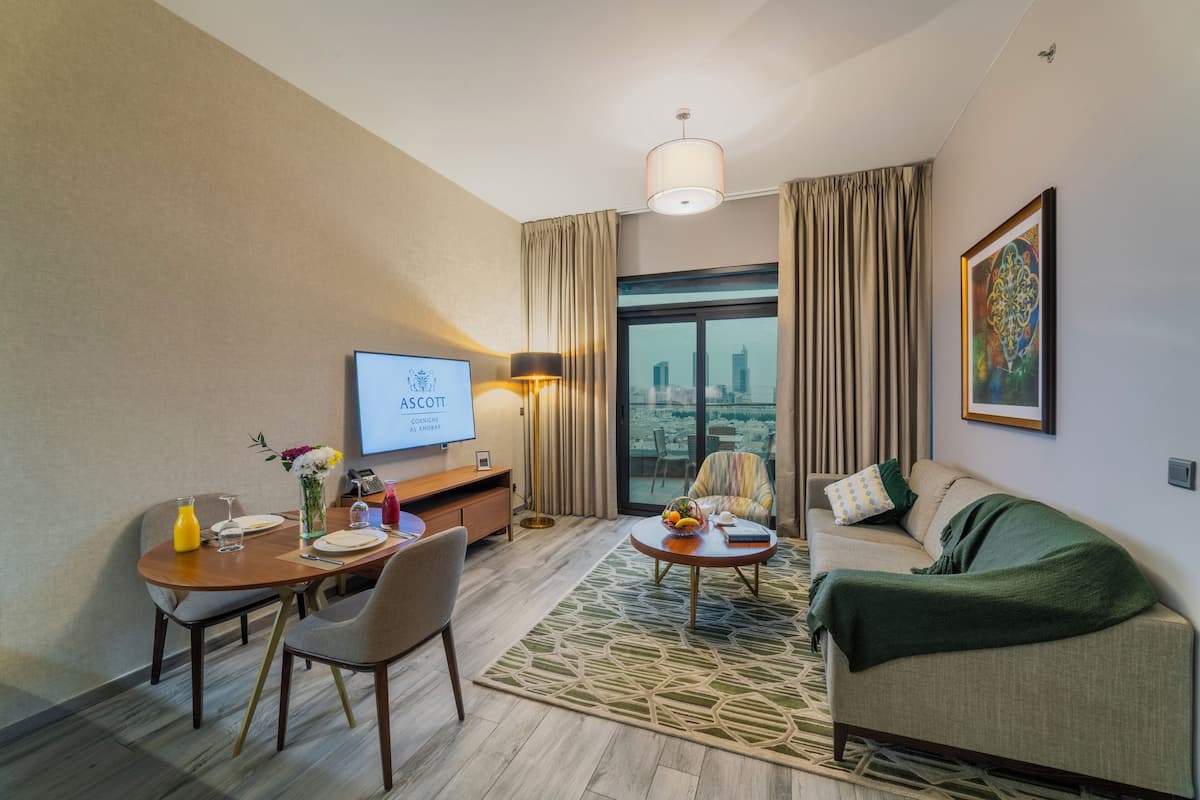 Habitación de hotel con zona de comedor, sofá, televisor y un amplio ventanal con vistas a la ciudad.