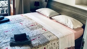 Un lit bien fait, recouvert d’une couvre-lit à motifs floraux, avec une tête de lit en bois et une table de chevet surmontée d’une lampe.