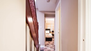 Un pasillo con una escalera de madera, una cama con colcha a cuadros y una mesita de noche con una lámpara.